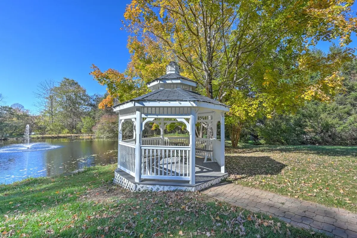A scenic view of the gazebo by a pond, surrounded by trees and a winding pathway. The tranquil setting offers versatile space for outdoor receptions, quiet chats, or a nightcap as the sun sets over the water.