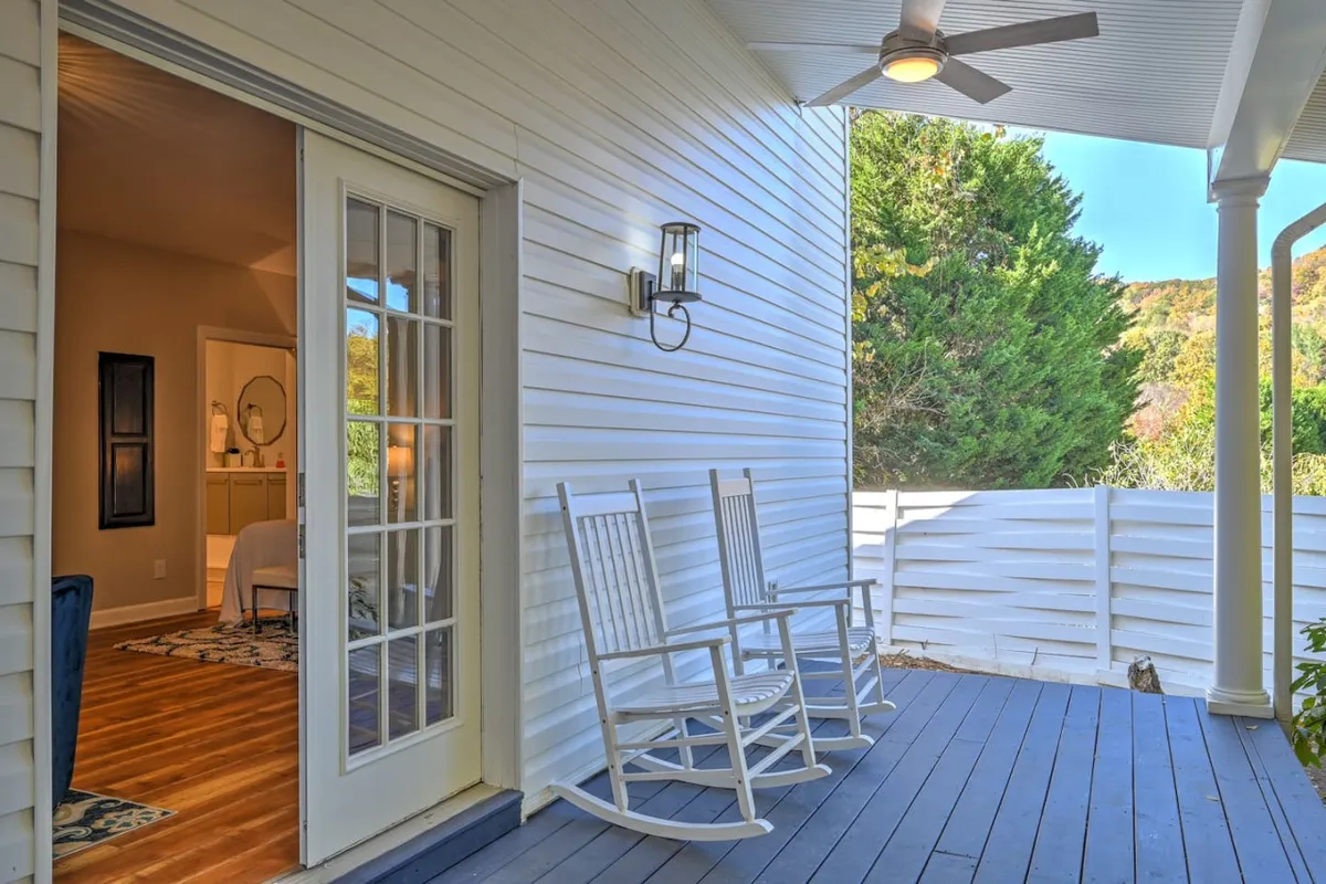 An inviting exterior porch scene featuring classic rocking chairs arranged just outside a doorway, with a soft outdoor light fixture and lush greenery in the background. The space is designed for relaxation, reading, or socializing while immersed in nature.