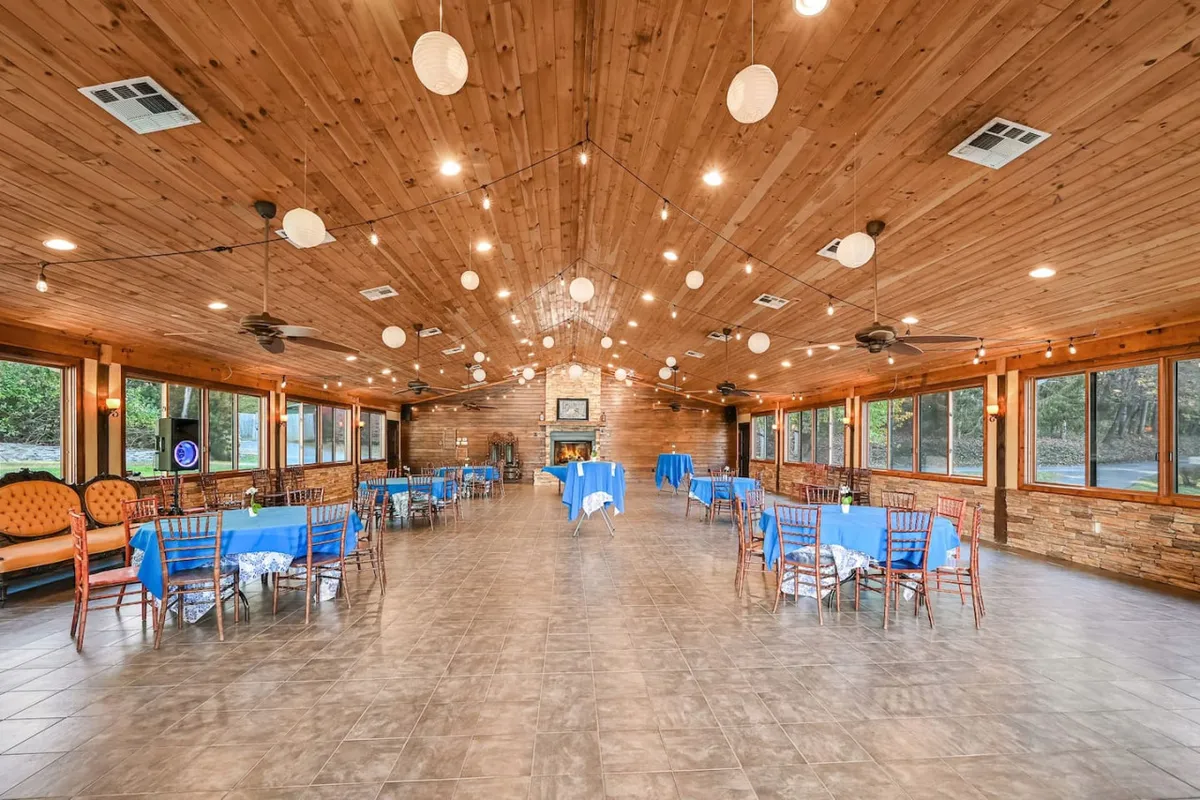 A spacious event center interior featuring a warm wooden ceiling, large windows that flood the space with natural light, decorative lights overhead, and several dining tables arranged for gatherings. The focal point is the openness and flexibility of the layout, ideal for hosting a variety of events from weddings to corporate functions.