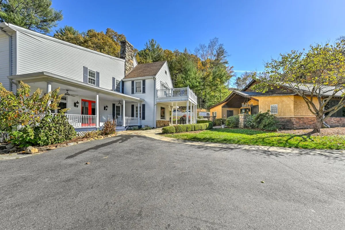 This image showcases the main house with its inviting red door, spacious outdoor porch, and surrounding trees. The scene includes both the main and guest house, and hints at the natural tranquility of the estate. The outdoor setup is perfect for family gatherings, outdoor dining, and relaxed social events, with picturesque sunset views from the porch and ample space for everyone to unwind together after a day exploring Asheville.