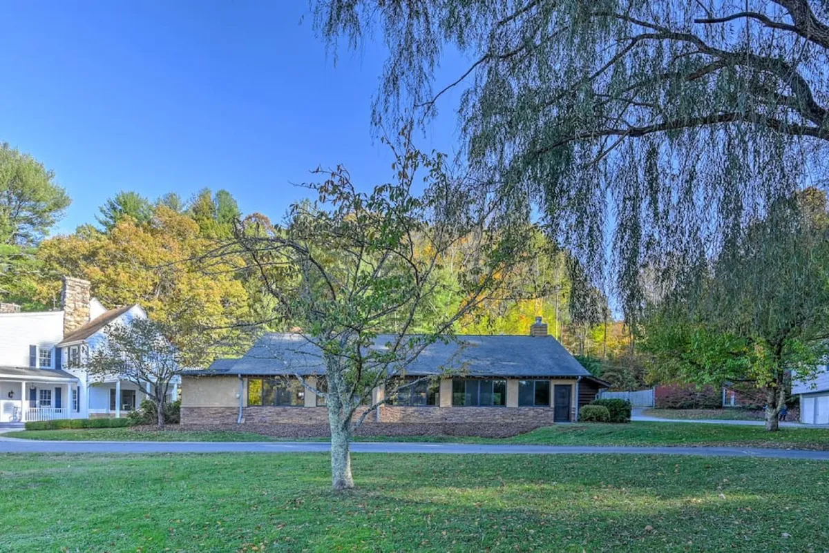 An exterior view of the home, framed by tall trees and a sweeping driveway, with a spacious yard suggesting ample room for outdoor gatherings and activities. The serene atmosphere is heightened by the natural greenery and open space.