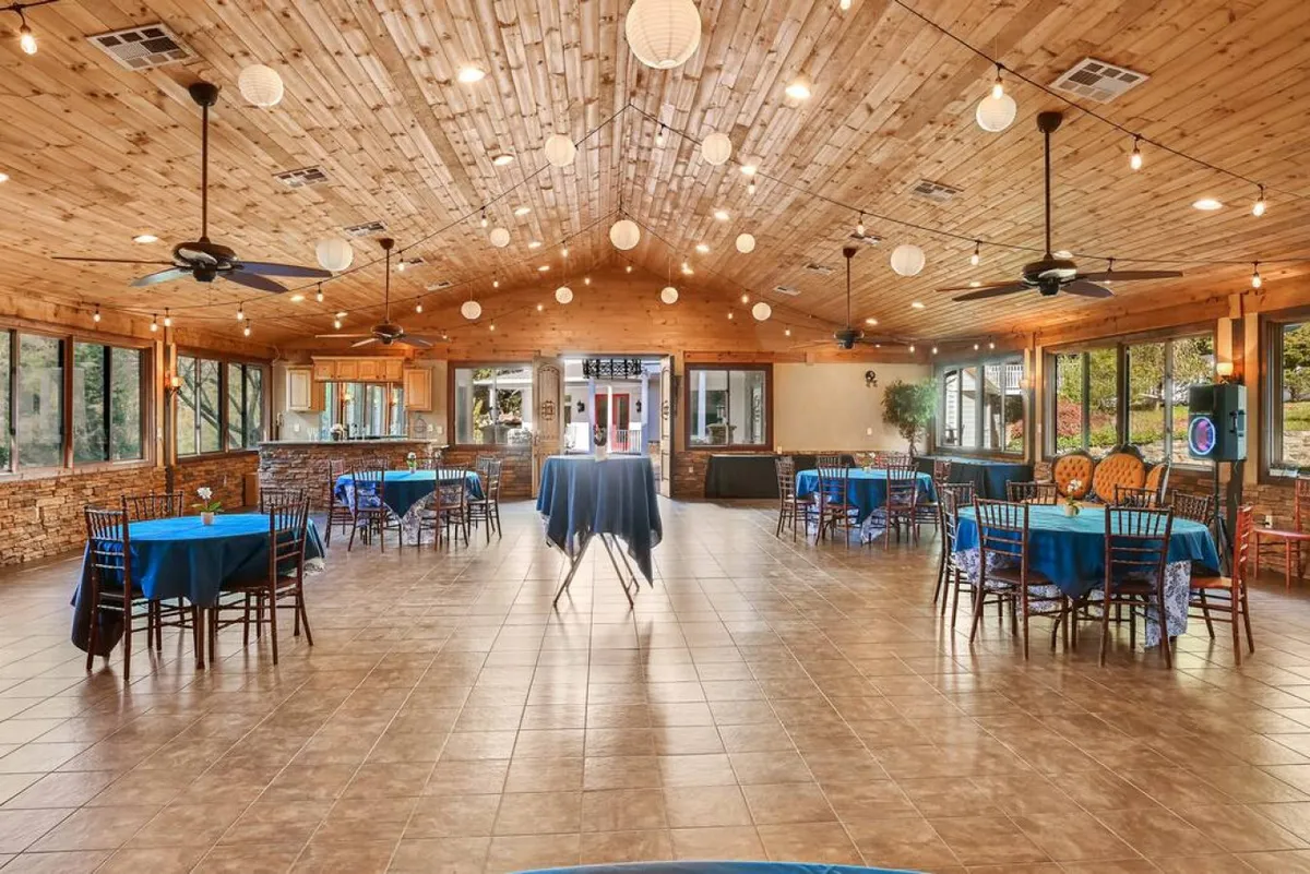 An inviting interior shot highlighting a large wooden ceiling, hanging lanterns, tables with blue tablecloths, and wide windows letting in natural light. The setup suggests a lively event, such as a wedding reception or corporate party, emphasizing the celebratory vibe and flexibility of the space.