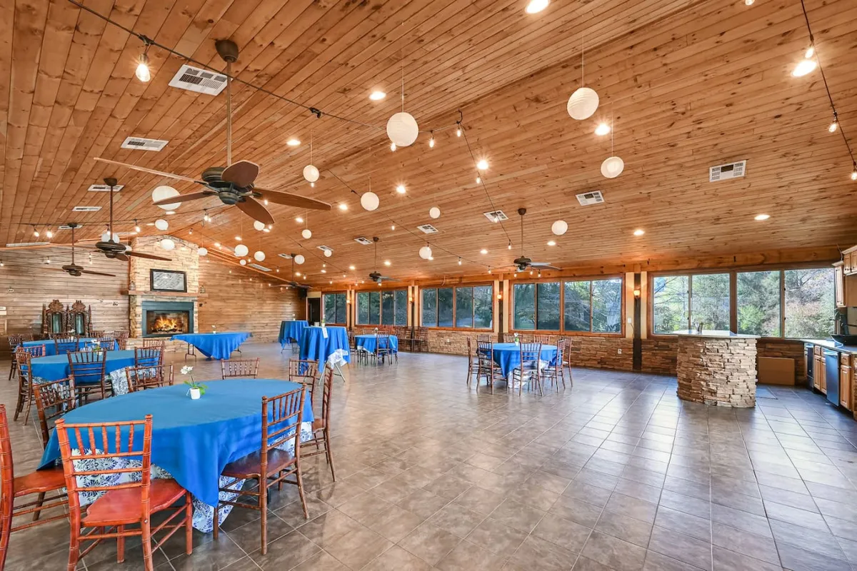 This vibrant interior image features a spacious event center with a wooden ceiling, large windows, decorative lanterns, blue tablecloths, and a cozy fireplace. The space is arranged for celebrations, suggesting both wedding receptions and corporate gatherings in a warm, communal setting.