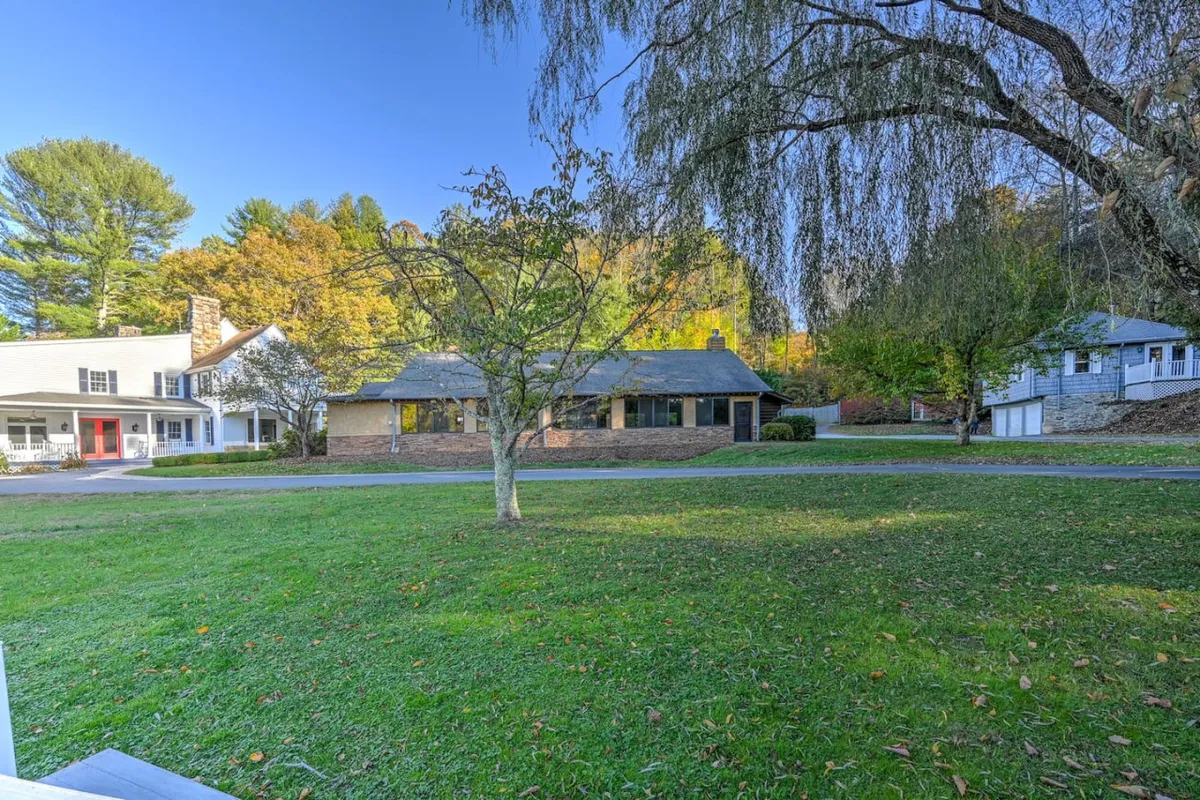 A broad exterior shot of the main house surrounded by mature trees and a sweeping driveway, with a lush lawn ideal for gatherings, lawn games, or picnics. The house is framed by the vibrant greenery of the landscape.
