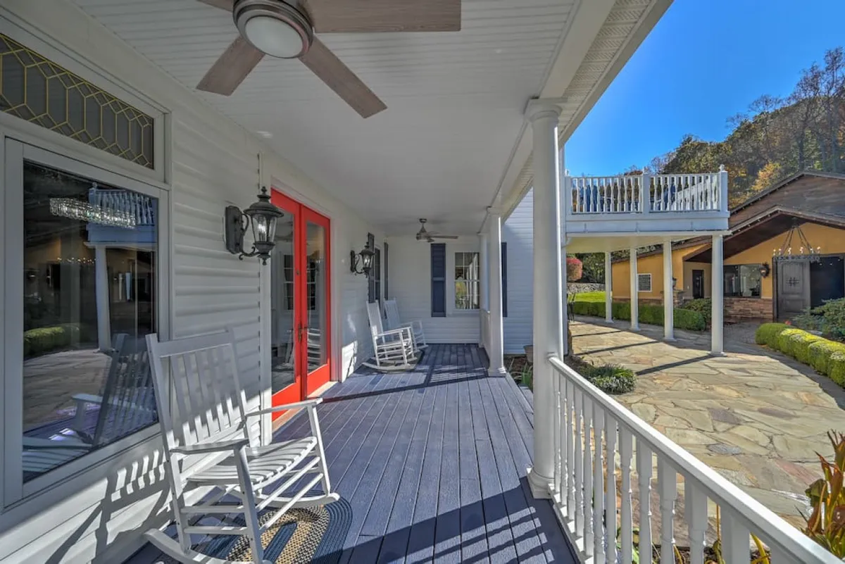 A welcoming porch scene with rocking chairs, a bright red door, an outdoor ceiling fan, and a stone pathway leading through surrounding greenery. The space offers multiple seating options for enjoying coffee, reading, or socializing in a serene, shaded spot.