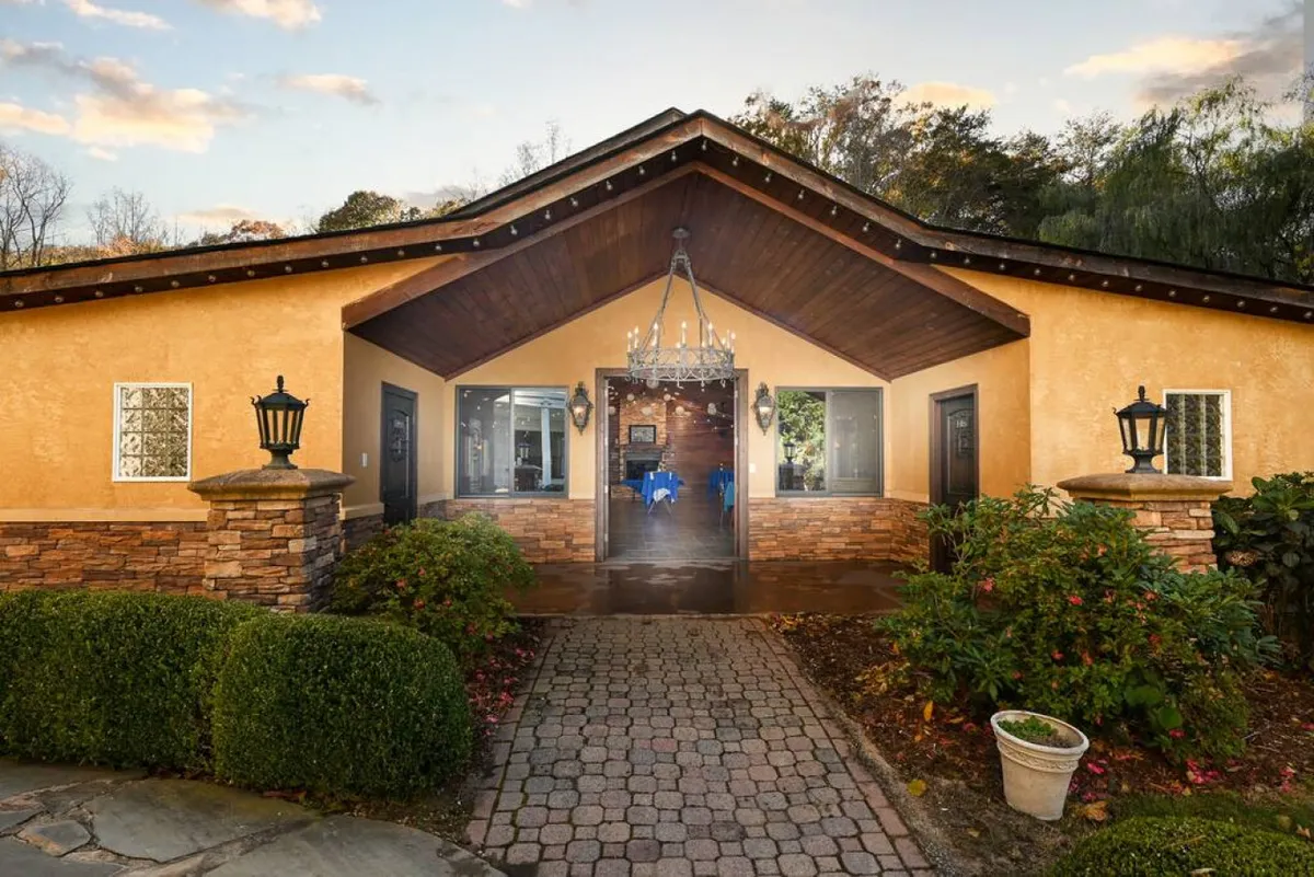 The photo features the elegant main entrance to the estate, with a chandelier illuminating the stonework and manicured landscaping. The scene is both grand and welcoming, suggesting a space designed for special occasions and group celebrations, while also offering a tranquil atmosphere for guests arriving after an eventful day.