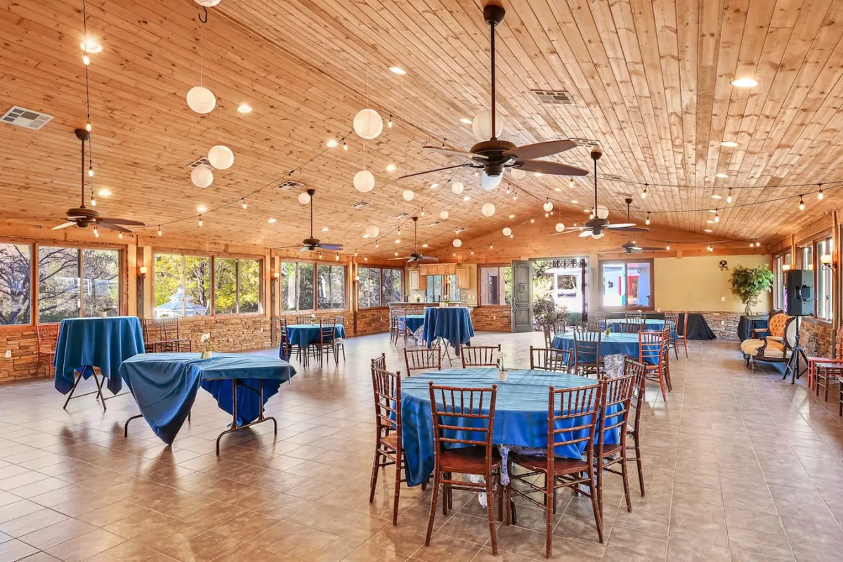 A view of the event center set with round tables dressed in blue tablecloths, large windows allowing sunlight to flood the space, decorative ceiling fans, and string lights. The arrangement highlights the social and festive nature of gatherings held here.