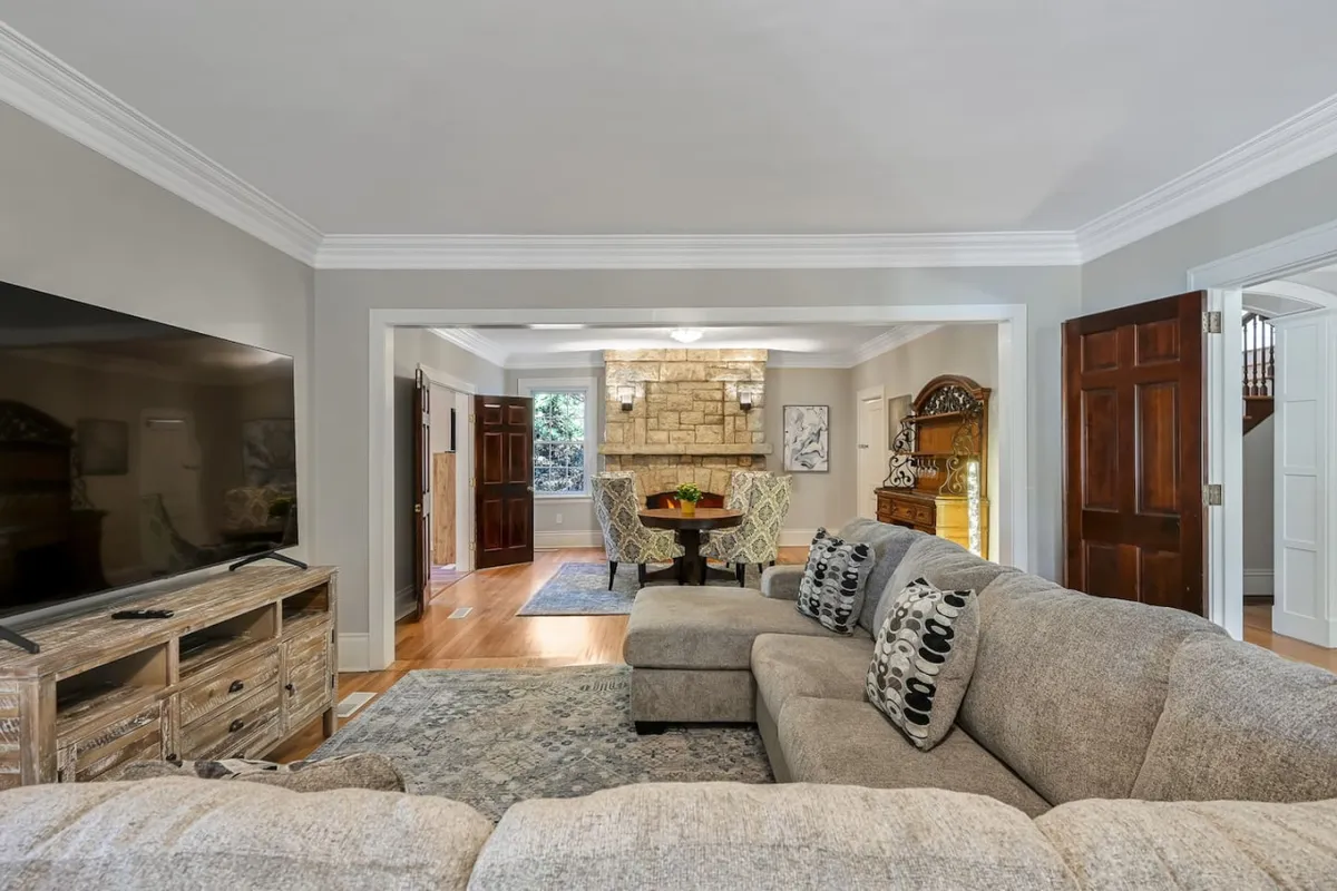 A spacious open-concept living and dining room featuring a large sectional sofa, a stone fireplace, a wooden dining table with chairs, and warm wooden doors. The arrangement encourages interaction, relaxation, and shared experiences, with the fireplace providing a cozy focal point for gatherings.