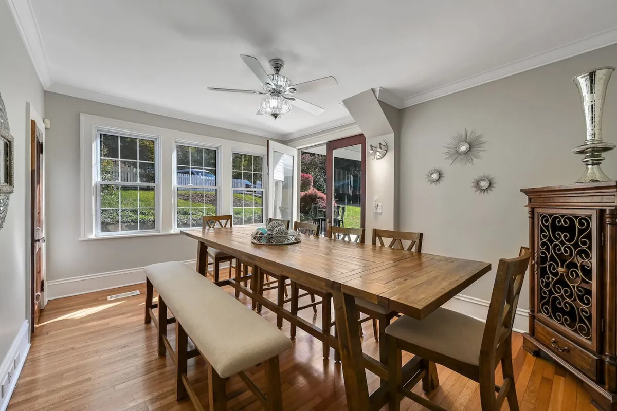 A long wooden dining table sits beside large windows that let in ample natural light. The space is decorated with a wooden sideboard and wall art, creating an inviting atmosphere for family meals and celebrations.